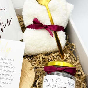 May include: A gift box with a white mug, a white washcloth tied with a burgundy ribbon, a gold spoon, a candle jar with a burgundy ribbon, and a card. The candle jar reads "Spark The Spirit of Godmother". The box is filled with brown crinkle paper.