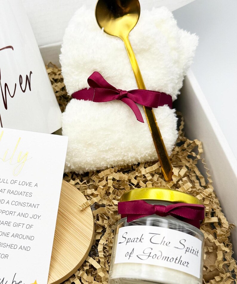 May include: A gift box with a white mug, a white washcloth tied with a burgundy ribbon, a gold spoon, a candle jar with a burgundy ribbon, and a card. The candle jar reads "Spark The Spirit of Godmother". The box is filled with brown crinkle paper.