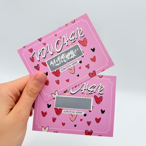 May include: Two pink scratch-off cards with a heart pattern. The top card says "You  Catcher" and the bottom card says "Cher" with a silver scratch-off area. Both cards say "Scratch Here".