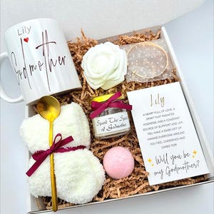 May include: A gift box with a white mug that says "Lily Godmother", a white rose, a gold bracelet, a small candle, a pink bath bomb, a gold spoon, and a card that says "Will you be my Godmother?"