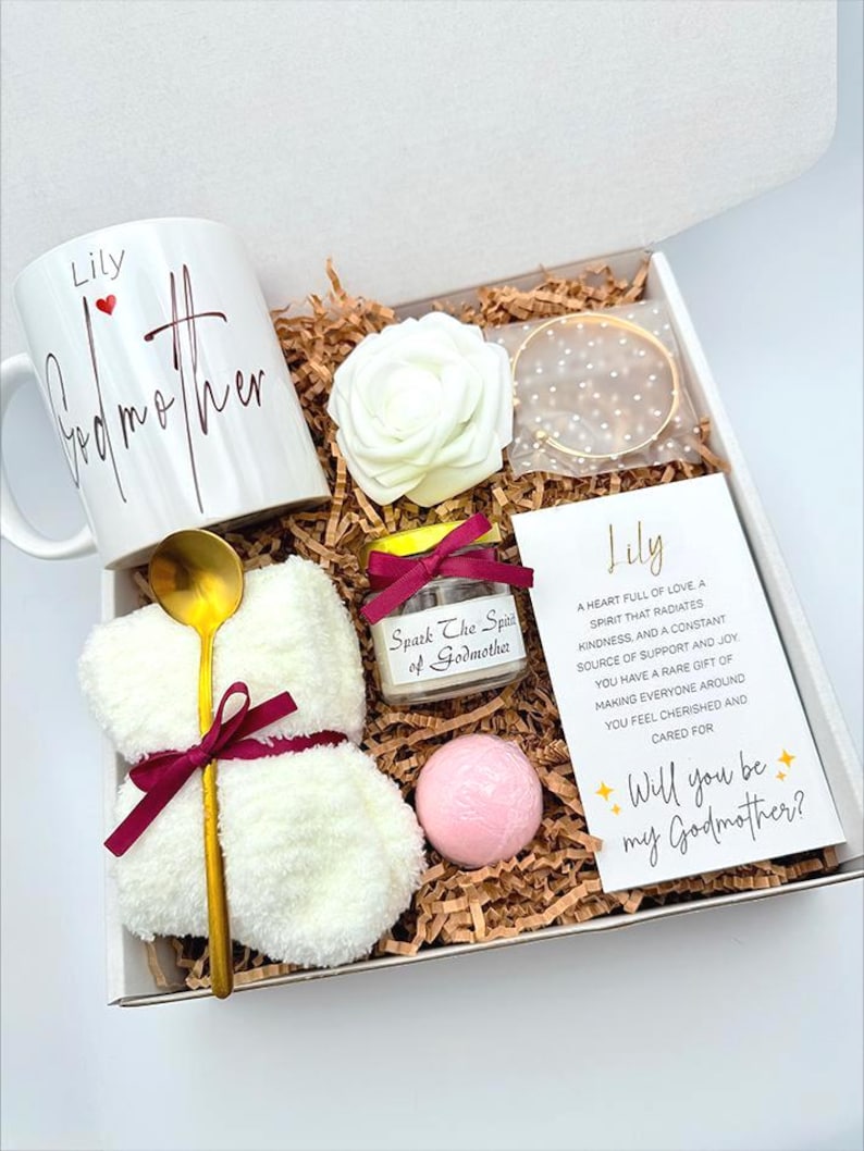 May include: A gift box with a white mug that says "Lily Godmother", a white rose, a gold bracelet, a small candle, a pink bath bomb, a gold spoon, and a card that says "Will you be my Godmother?"