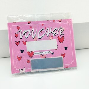 May include: Pink scratch-off voucher with a heart pattern and the word "VOUCHER" in white lettering. The voucher has a silver scratch-off area with the text "SCRATCH HERE" above it.