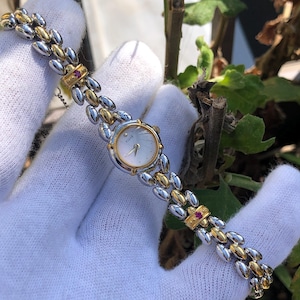 May include: A two-tone wristwatch with a silver and gold band. The watch face is round with a light-coloured background and gold-coloured hands. The band is made of oval-shaped links. A small red stone is set into the band near the watch face.