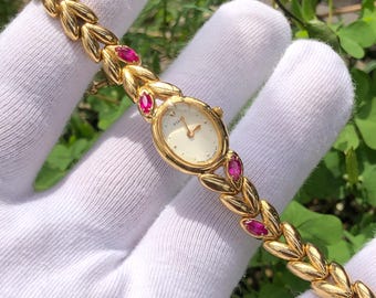 May include: A gold-toned wristwatch with a cream-colored face and ruby accents. The watch features an oval face and a delicate bracelet-style band with leaf-like details. The watch is a classic and elegant piece of jewelry.