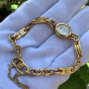 Puede incluir: Un reloj de pulsera dorado con una esfera ovalada y una pulsera de eslabones. La esfera del reloj es de color claro con números y manecillas negras. La pulsera presenta una mezcla de eslabones de cadena y barras rectangulares. Pequeñas gemas rojas acentúan la esfera.