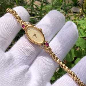 May include: A gold Seiko wristwatch with an oval face and a braided band. The watch has a cream dial with gold hands and markers. It is decorated with red and clear gemstones on the sides of the watch face.