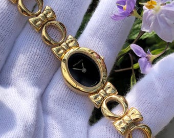 May include: A gold-toned Nina Ricci wristwatch with a black oval face and a small diamond accent. The watch features a bow-shaped link bracelet. The watch is displayed on a white surface, with purple flowers in the background.