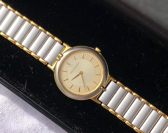 May include: A gold-toned wristwatch with a cream-colored face and a white and gold band. The watch face features the Yves Saint Laurent logo. The band is composed of rectangular links. The watch is displayed in a black box.
