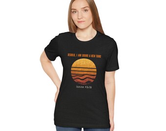 Può includere: T-shirt nero con un design a tramonto e il testo "BEHOLD, I AM DOING A NEW THING" e "Isaiah 43:19". Il design è un cerchio con linee orizzontali arancioni e gialle. La maglietta è realizzata in materiale morbido.