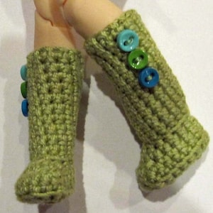 Może przedstawiać: Para zielonych, szydełkowych getrów, każdy ozdobiony trzema kolorowymi guzikami w odcieniach niebieskiego i zielonego. Ręcznie robione getry mają teksturowany wzór. Artykuł jest prawdopodobnie dla lalki lub podobnego.