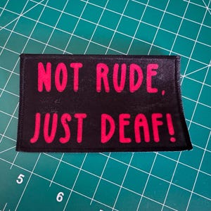 May include: A rectangular black fabric patch with a stitched border featuring the bright pink text NOT RUDE, JUST DEAF! in a bold, sans-serif font. The patch is positioned on a green cutting mat with visible white grid lines and numbers.