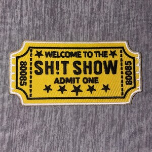 May include: Yellow patch with black text that reads "WELCOME TO THE SH!T SHOW ADMIT ONE" with four stars. The patch has a white border and the numbers "80085" on each side. The patch is rectangular with a ticket-like shape.