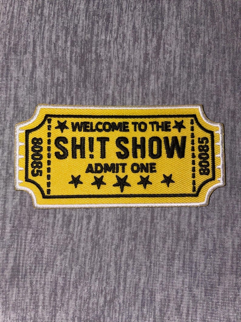 May include: Yellow patch with black text that reads "WELCOME TO THE SH!T SHOW ADMIT ONE" with four stars. The patch has a white border and the numbers "80085" on each side. The patch is rectangular with a ticket-like shape.