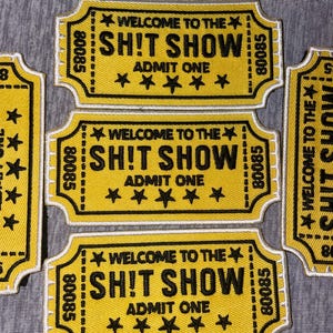May include: Yellow embroidered patches with black text that reads "WELCOME TO THE SH!T SHOW ADMIT ONE" and five black stars. The patches have a ticket shape with a white border and the number "80085".