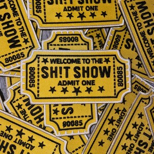 May include: A collection of yellow and black fabric patches. Each patch is shaped like a vintage ticket and features the text "WELCOME TO THE SH!T SHOW" and "ADMIT ONE". The patches have a star design and the number "80085".