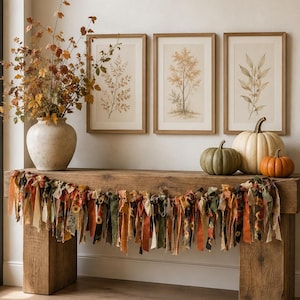 May include: A rustic wooden console table features a fabric rag garland in autumn colours of orange, brown, and green. A ceramic vase holds dried fall foliage, while three framed botanical prints hang above. Three pumpkins sit on the table surface.