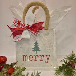 Könnte beinhalten: Eine beige Geschenk-Tasche aus Jute mit geflochtenem Griff, verziert mit einer rot-weißen Schleife und Seidenpapier. Die Tasche zeigt einen grünen Weihnachtsbaum und das Wort "merry" in Rot. Festliche Weihnachtsdekoration.