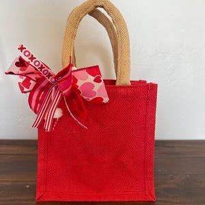 May include: A small red burlap tote bag with a brown jute handle. The bag is decorated with a pink and red heart patterned ribbon bow with the text "XOXOXOXO" printed on it.