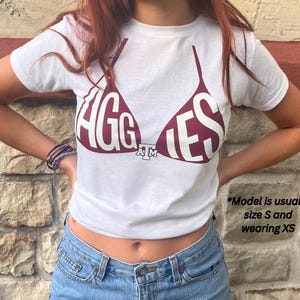 May include: White t-shirt with a maroon bra graphic that says "AGGIES". The shirt is short-sleeved and made of a soft material. The shirt is paired with blue jeans.