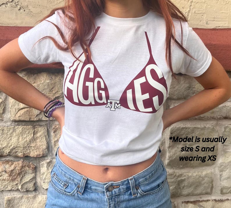 May include: White t-shirt with a maroon bra graphic that says "AGGIES". The shirt is short-sleeved and made of a soft material. The shirt is paired with blue jeans.