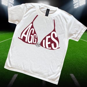 May include: White t-shirt with a maroon bra graphic. The bra design spells out "AGGIES" in white letters. A small "A&M" logo is centered below the bra. The shirt is on a green field background.