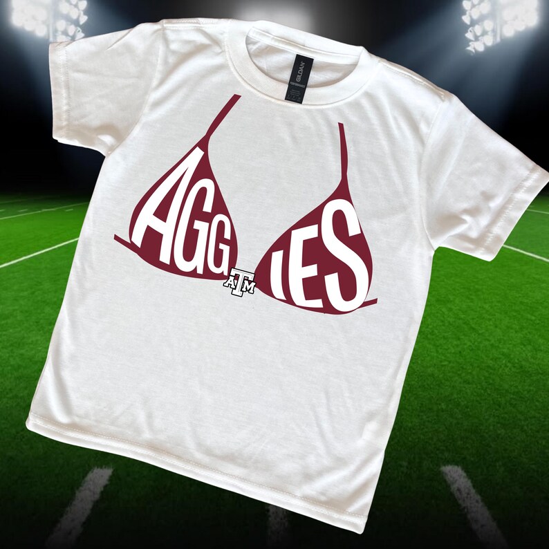May include: White t-shirt with a maroon bra graphic. The bra design spells out "AGGIES" in white letters. A small "A&M" logo is centered below the bra. The shirt is on a green field background.
