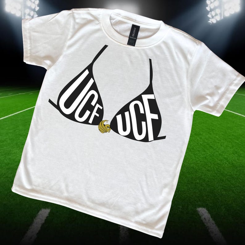 May include: White t-shirt featuring a black bra design with the letters "UCF" in bold white font. A gold emblem is centered between the bra cups. The shirt is displayed on a green field background.