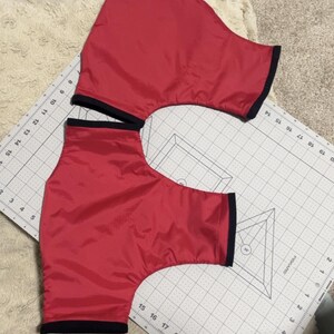 May include: Two red fabric pieces with black trim, likely for a sewing project. The fabric is laid out on a white gridded surface with markings in inches.