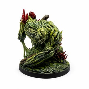 May include: A detailed miniature figure of a swamp creature, predominantly green with red accents. The figure has a textured, organic appearance, mounted on a black circular base. The creature appears to be a fantasy or horror-themed character.