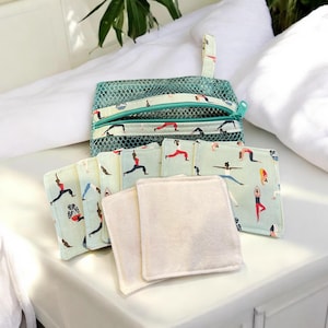May include: A set of reusable makeup remover pads in a green mesh bag. The pads are made of soft, natural fabric and feature a pattern of women doing yoga poses. The bag has a zipper closure.