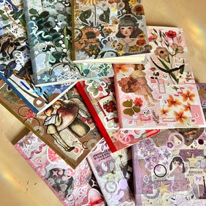 May include: A selection of notebooks with decorative covers. The designs include floral patterns, vintage illustrations, and stickers. The covers feature sunflowers, butterflies, and images of girls, creating a collage effect with a vintage aesthetic.