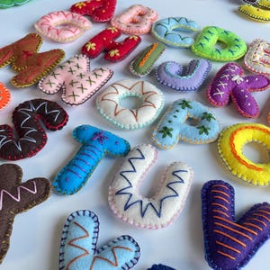 May include: A selection of colourful, felt letters and shapes, including the letters A, T, U, and V. The letters are in various colours, such as blue, pink, and yellow, with embroidered details. The shapes include hearts and donuts.