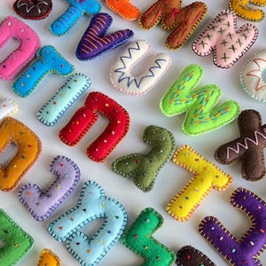 May include: A collection of colorful, felt letters in various colors, including pink, blue, green, and yellow. Each letter is hand-stitched with contrasting thread and embellished with small, colorful beads. The letters are arranged on a white surface.