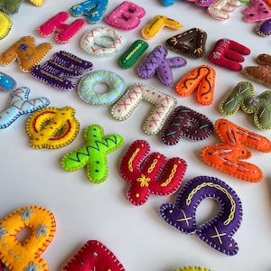 May include: A selection of brightly coloured felt letters, including pink, orange, yellow, and purple. Each letter is hand-stitched with contrasting thread, giving a textured look. The letters are arranged on a white surface.