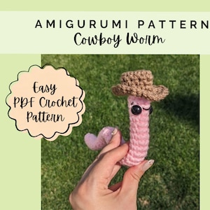 May include: A pink crocheted worm toy wearing a small brown crocheted cowboy hat with black plastic eyes. The image includes the text AMIGURUMI PATTERN Cowboy Worm and Easy PDF Crochet Pattern. The worm has a segmented body and a curved tail.