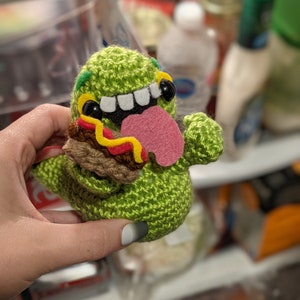 May include: Green crocheted plush toy with a wide open mouth, white teeth, and a pink tongue. The toy is holding a brown crocheted hot dog with yellow mustard.