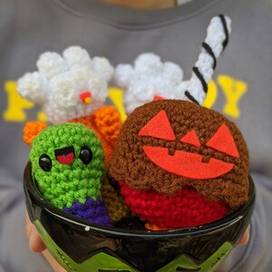 Może przedstawiać: Zielona ceramiczna miska w kształcie twarzy Frankensteina zawiera kolekcję postaci z Halloween wykonanych na szydełku. Postacie obejmują dynię, ducha, kukurydzę cukrową i zielonego potwora.