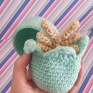 May include: A crocheted light blue egg with a crocheted tan dinosaur inside. The egg is open and the dinosaur is peeking out.