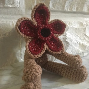 May include: A crocheted plush toy shaped like the monster from the show Stranger Things. The monster has a red flower-like head with a black center and brown legs.