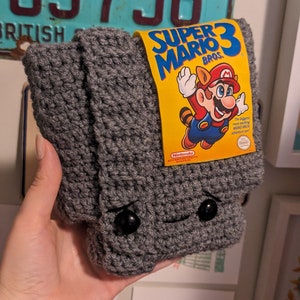 May include: A gray crocheted pouch featuring two black button eyes and a small stitched mouth. A yellow rectangular label is attached to the top, displaying the text Super Mario Bros. 3 with an illustration of Mario in a raccoon suit.