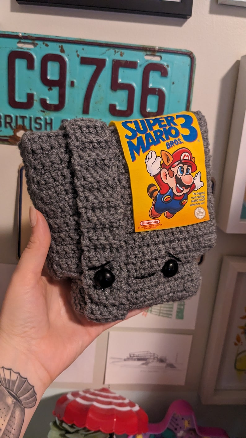 May include: A gray crocheted pouch featuring two black button eyes and a small stitched mouth. A yellow rectangular label is attached to the top, displaying the text Super Mario Bros. 3 with an illustration of Mario in a raccoon suit.