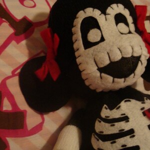 May include: A black and white plush toy with a skull face and a red bow. The toy is wearing a black and white skeleton costume with the word "Cute" on the chest.