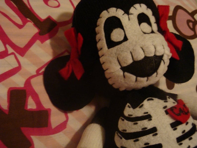 May include: A black and white plush toy with a skull face and a red bow. The toy is wearing a black and white skeleton costume with the word "Cute" on the chest.