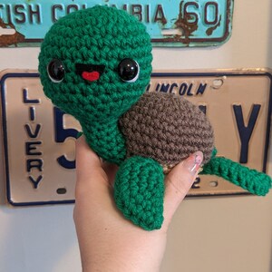 May include: A crocheted green turtle with brown shell and black eyes. The turtle has a red mouth and is smiling.