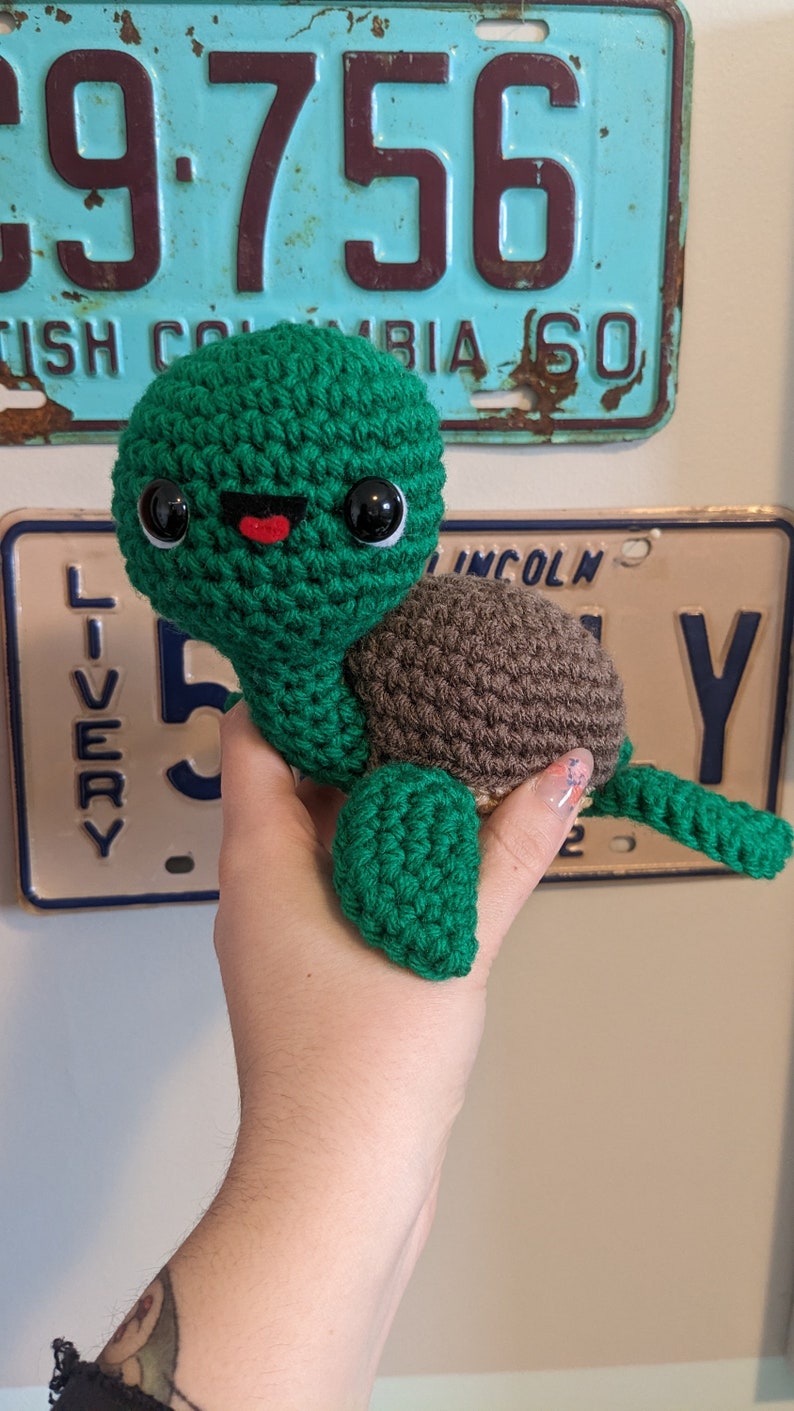 May include: A crocheted green turtle with brown shell and black eyes. The turtle has a red mouth and is smiling.