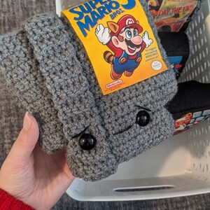 May include: A grey crocheted sleeve featuring two black circular buttons for eyes, with a yellow Super Mario Bros. 3 game label attached to the front. The sleeve is textured with a repeating stitch pattern and is held in a white plastic basket.