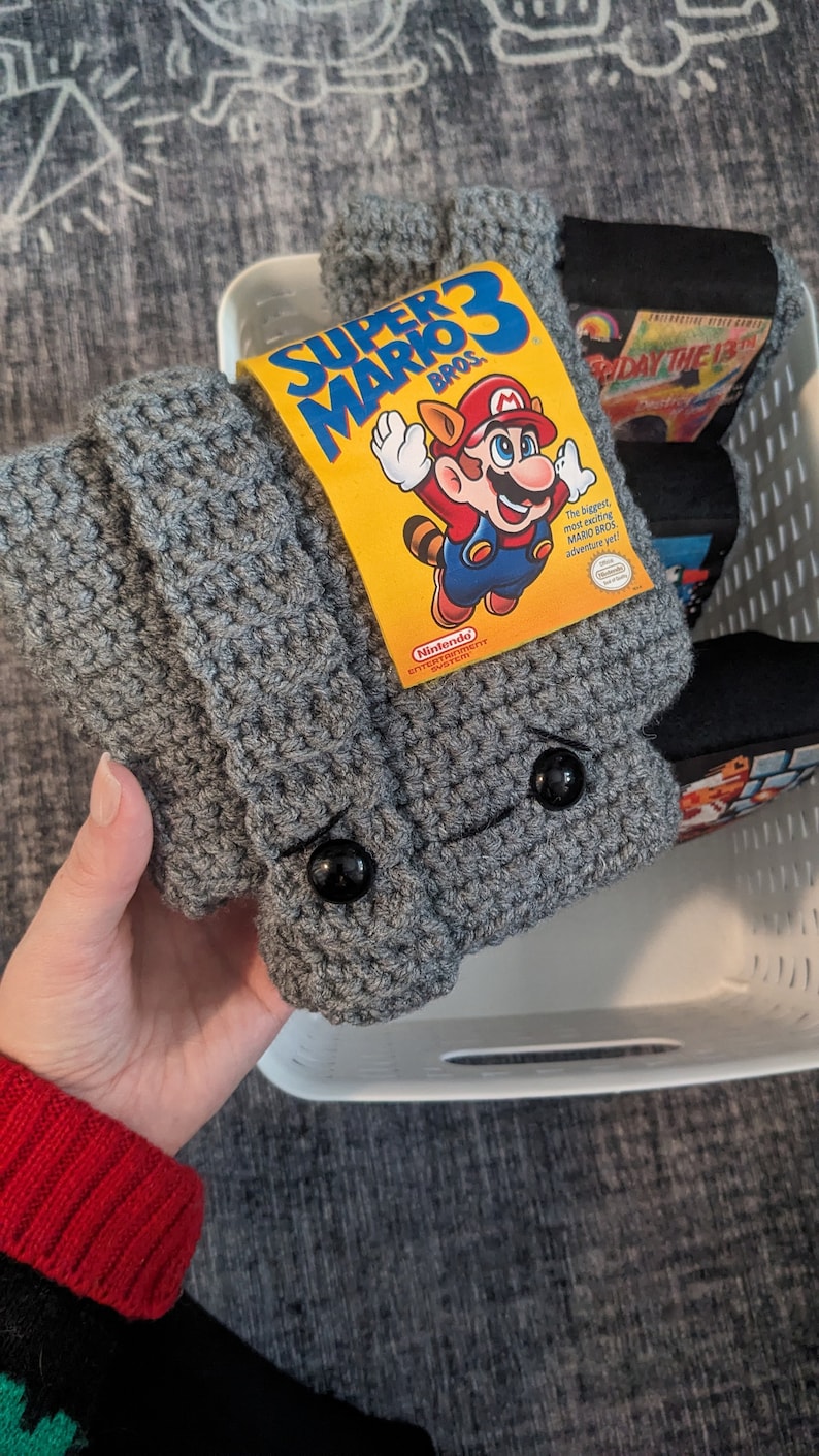 May include: A grey crocheted sleeve featuring two black circular buttons for eyes, with a yellow Super Mario Bros. 3 game label attached to the front. The sleeve is textured with a repeating stitch pattern and is held in a white plastic basket.