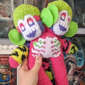 May include: A handmade plush toy with a pink body, green ears, and a white skeleton design. The toy has two heads, one with a purple face and the other with a red face. Both heads have white eyes and a stitched smile.