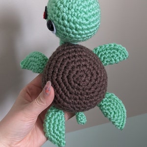 May include: A crocheted green sea turtle with brown shell and black eyes. The turtle is held in a hand.