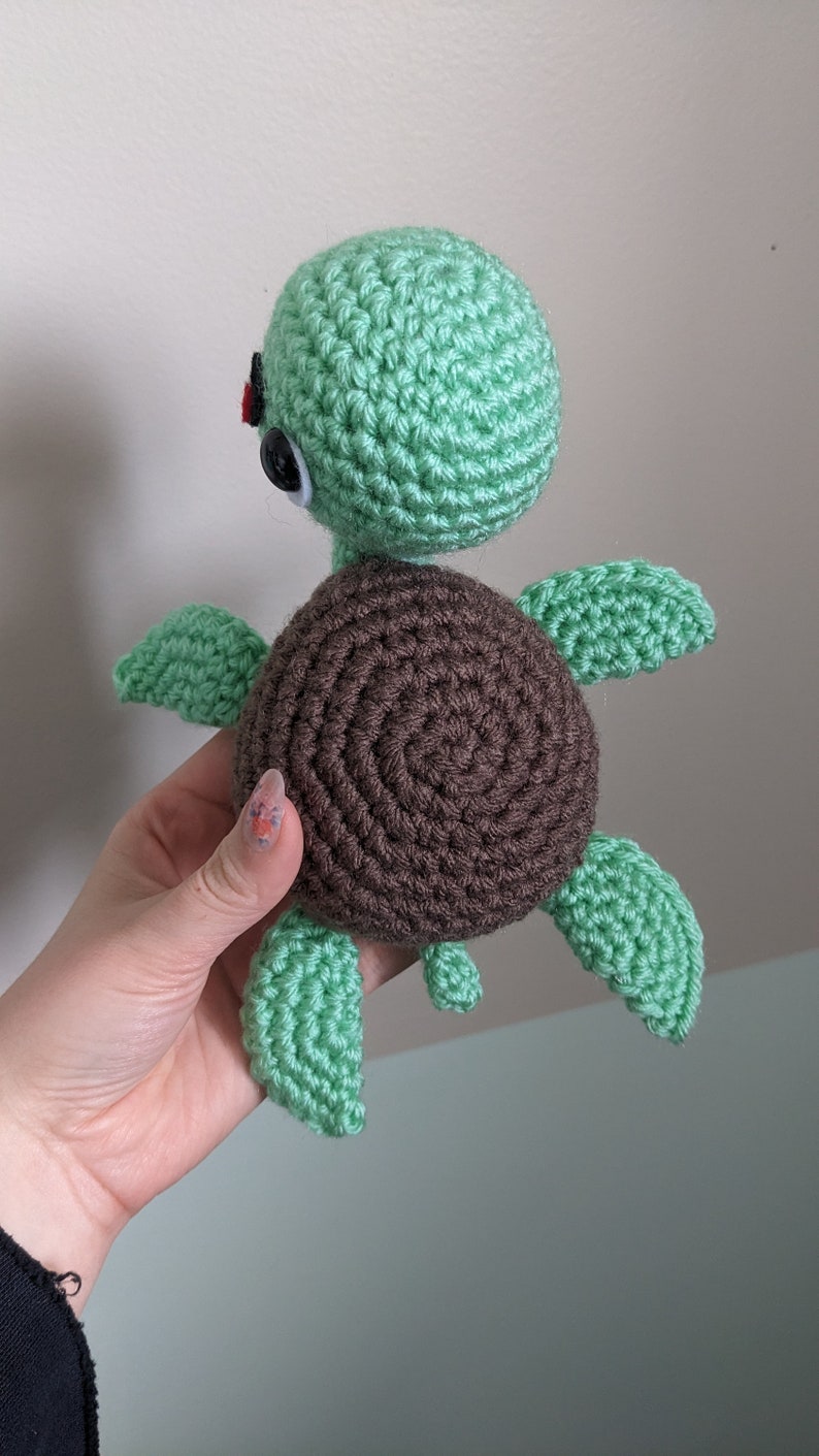 May include: A crocheted green sea turtle with brown shell and black eyes. The turtle is held in a hand.